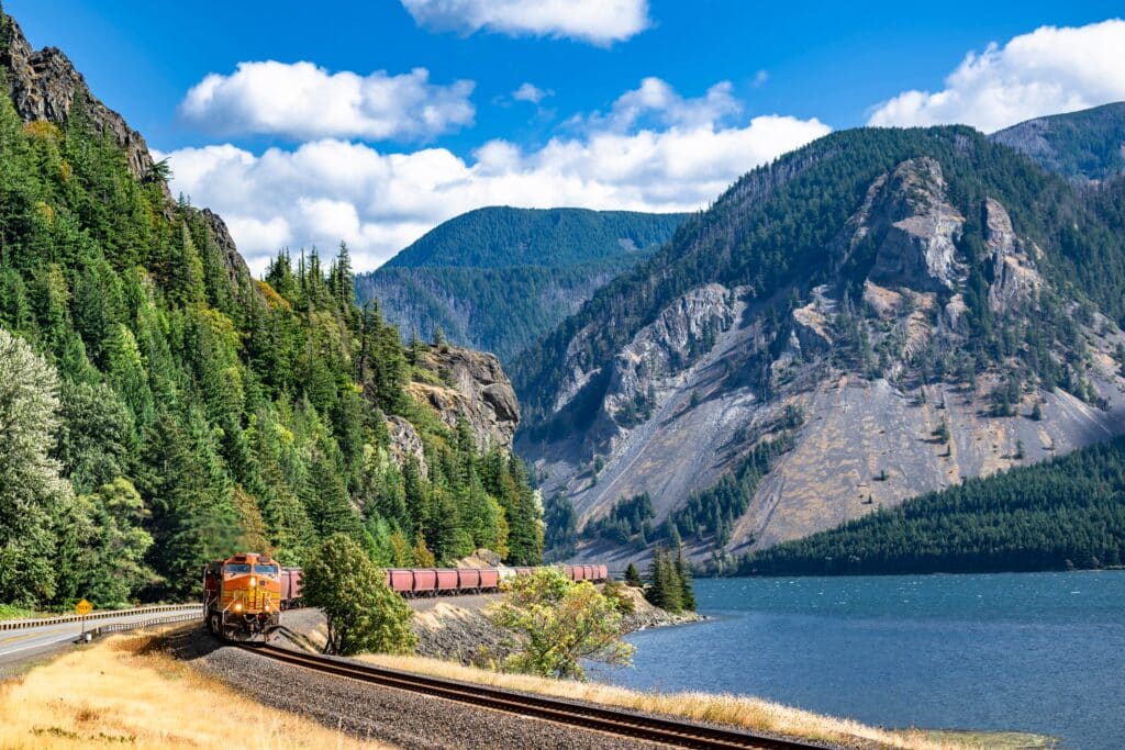 Landscape view of the locomotive pulls a train of freight cargo wagons on a railroad along Columbia River in Columbia Gorge scenic area with mountain ranges with evergreen trees along the river