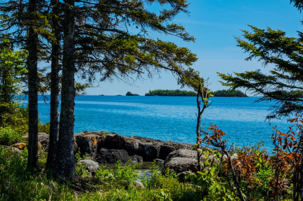 A view through the forest shows several of the tiny islands off the main island of Isle Royale NP in Lake Superior off Copper Harbor, MI. Large boulders surround a little water pool on shoreline.