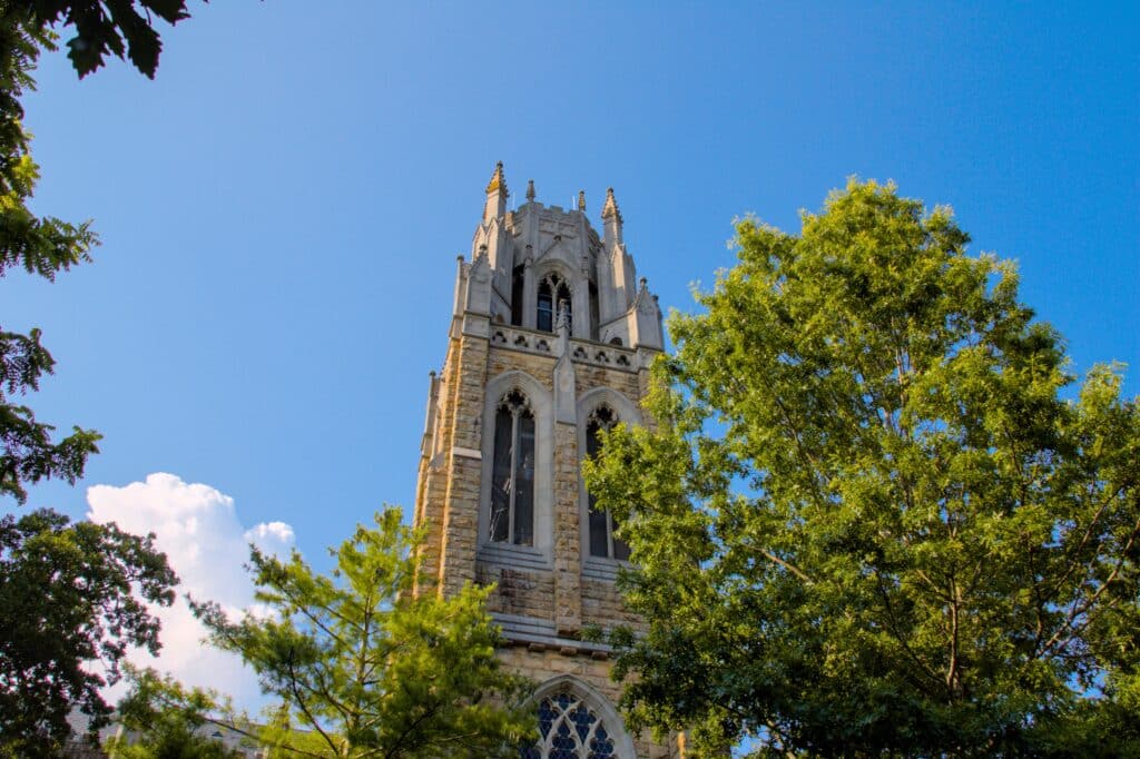 Gothic-style stone bell tower of All Saints’ Chapel at the University of the South in Sewanee, Tennessee, rises dramatically against a clear blue sky. Framed by lush green trees, the architecture feat