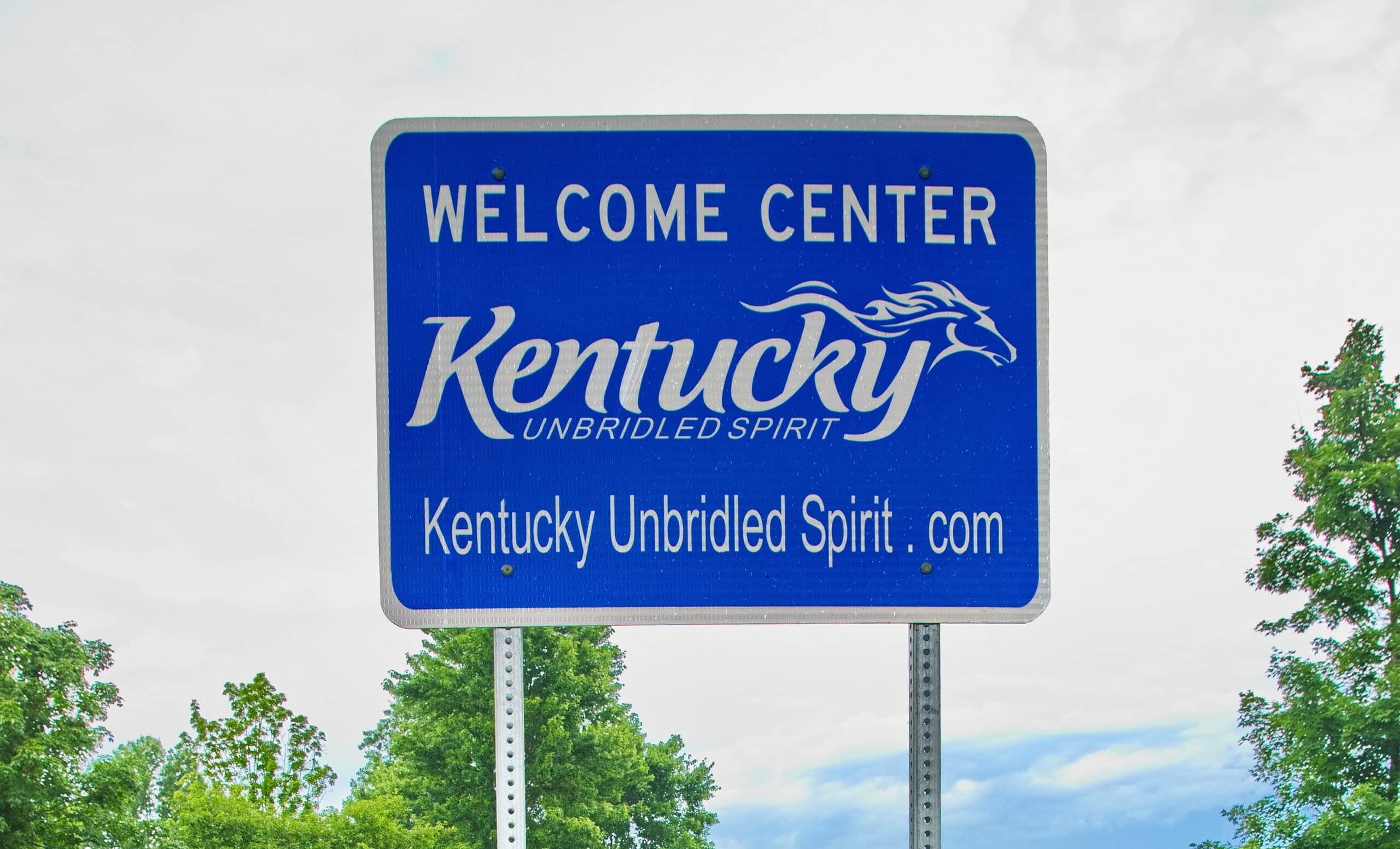 Kentucky Welcome Center highway sign featuring the state slogan Kentucky Unbridled Spirit against a cloudy sky and green trees, symbolizing travel and tourism.
