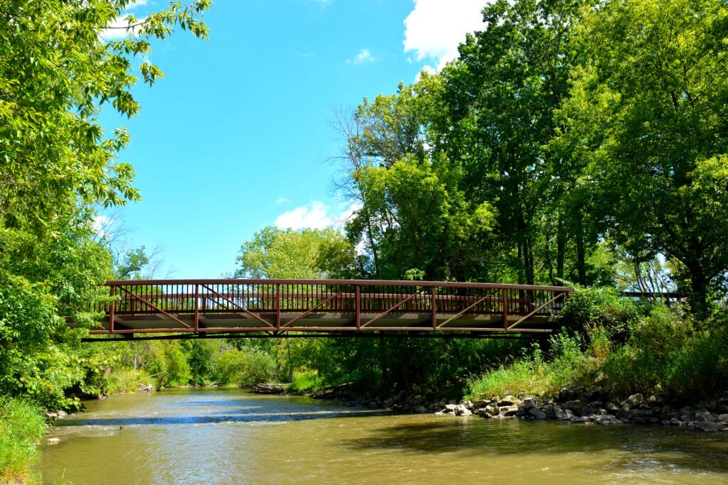 A foot bridge crossing Root River in Lincoln Park in Racine Wisconsin on a sunny summer afternoon.  The river flows under the bridge into the distance.  A few clouds highlight the blue sky.