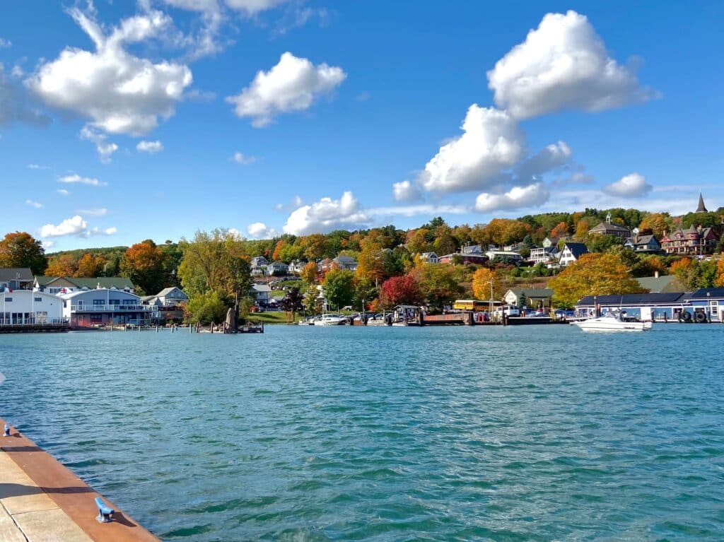 A stunning autumn landscape captured near Bayfield, Wisconsin, where vibrant fall foliage in shades of red, orange, and yellow contrasts beautifully against the deep blue-green waters of Lake Superior