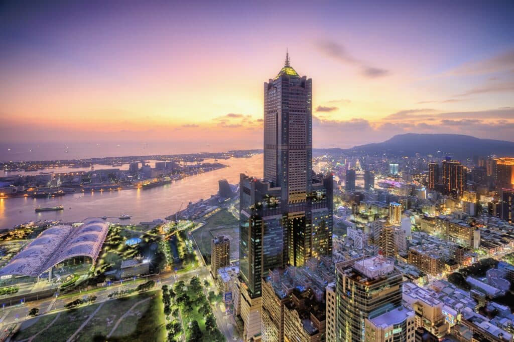 This magnificent aerial photograph captures the urban charm of Kaohsiung, Taiwan. The iconic 85 Sky Tower stands at the center, adjacent to the uniquely shaped Kaohsiung Exhibition Center and the glittering Kaohsiung Harbor. The afterglow of the sunset paints the sky in shades of pink, purple, and orange as the city lights begin to twinkle, perfectly capturing the prosperity and serenity of this port city at dusk. Ideal for travel guides, real estate development, Asian city studies, and high-end commercial backgrounds.