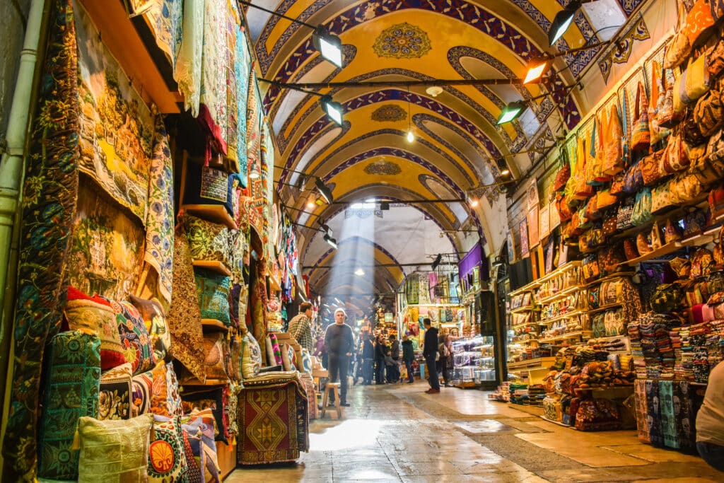 Istanbul, Turkey: People shopping in the Grand Bazar, handmade pillows, bags and carpets are on the wall for sale. The sunlight comes to inside from roof window.